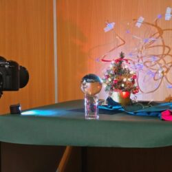 Een kleine fotoset op een tafel met een mini-kerstboompje, lampjes en een glazen bol op een standaard. Links staat een camera op statief gericht op de opstelling. Op de achtergrond zijn houten panelen en lichtsnoeren zichtbaar, vastgezet met tape.